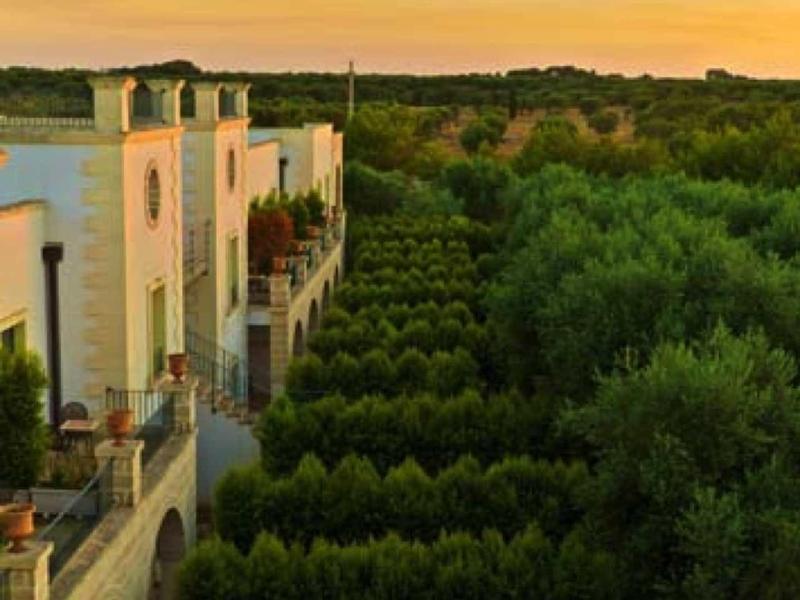 Ein helles Haus mit Balkon neben einem dichten, grünen Garten bei Sonnenuntergang.