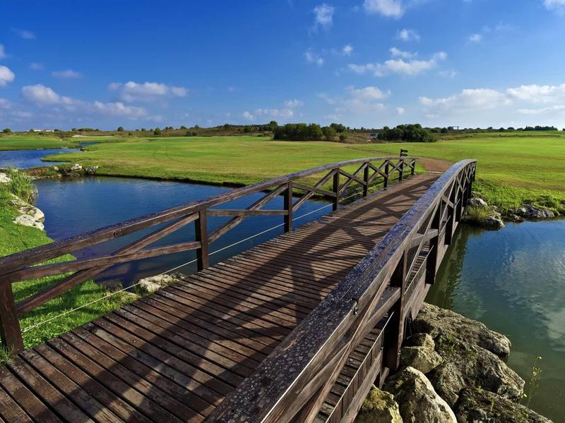 Holzbrücke über ruhigen Fluss, umgeben von grünen Wiesen und blauem Himmel mit Wolken.