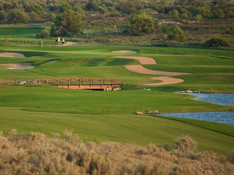 Grüner Golfplatz mit Brücke, Sandbunkern und Wasserfläche vor bewaldetem Hügel.