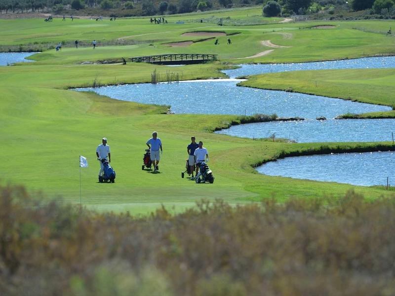 Drei Personen spielen Golf auf grüner Wiese mit mehreren Wasserhindernissen bei sonnigem Wetter.