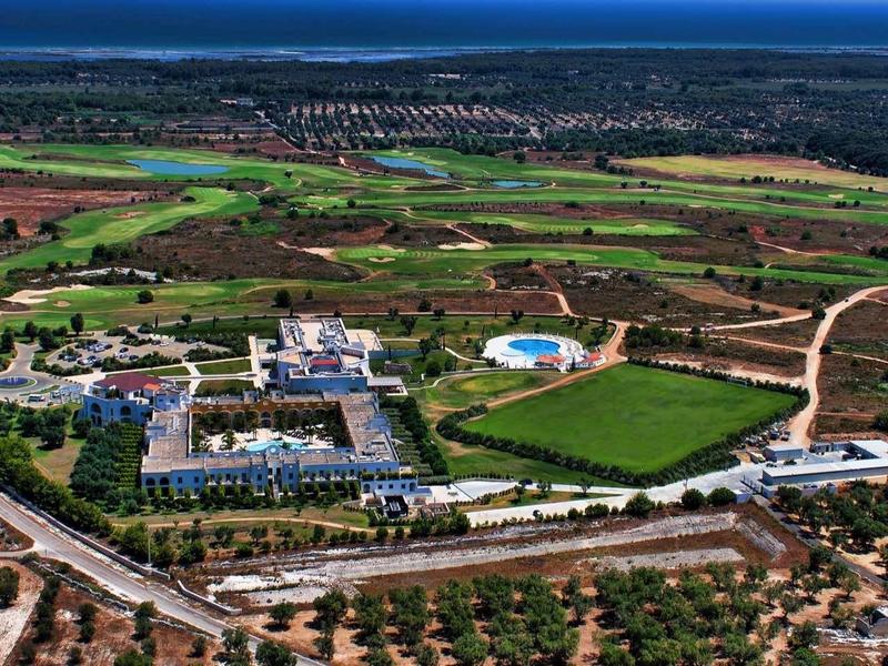 Landschaft mit Feldern, mehreren Gebäuden und einem großen grünen Sportplatz mit blauem Schwimmbecken.