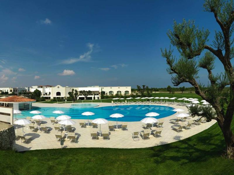 Großer Pool vor weißen Gebäuden, blaue Liegestühle und Sonnenschirme, grüner Baum rechts, blauer Himmel.