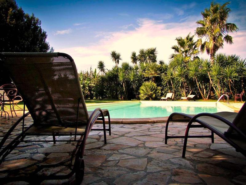 Pool mit blauem Wasser, umgeben von Palmen, zwei Sonnenliegen auf gepflastertem Boden.