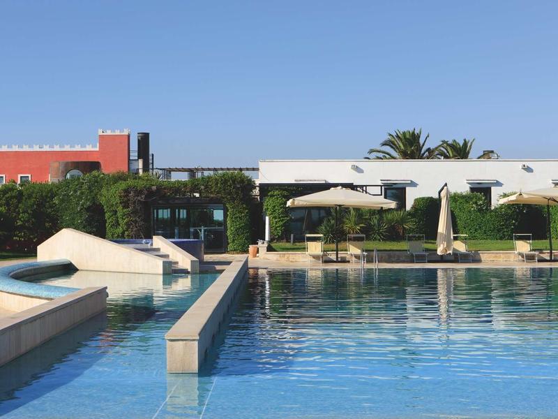 Área moderna de piscina con agua azul clara, tumbonas y sombrillas bajo un cielo azul.