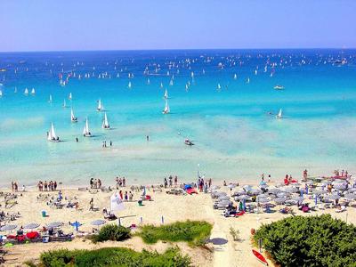 Strand mit Sonnenschirmen, Menschen und zahlreichen Segelbooten im türkisblauen Meer.