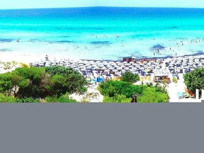 Breiter Sandstrand mit blauer Meeresbucht, vielen Sonnenschirmen und grünen Hügeln im Hintergrund.