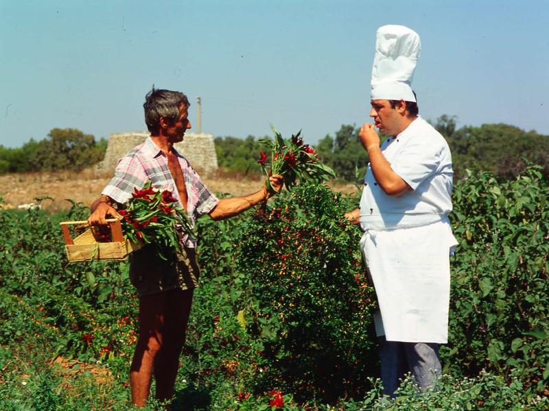 Zwei Männer stehen im Feld, einer trägt Kochmütze, der andere trägt Korb und Zweige.