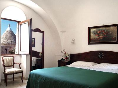 Slaapkamer met groen bed, houten stoel en open raam met uitzicht op een trullitoren.