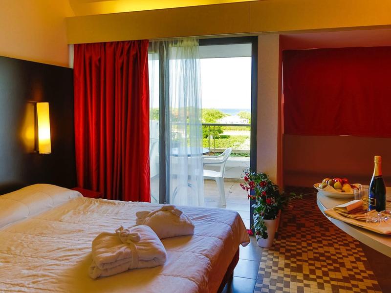 Chambre d'hôtel lumineuse avec lit double, rideau rouge et balcon avec vue sur le paysage verdoyant.