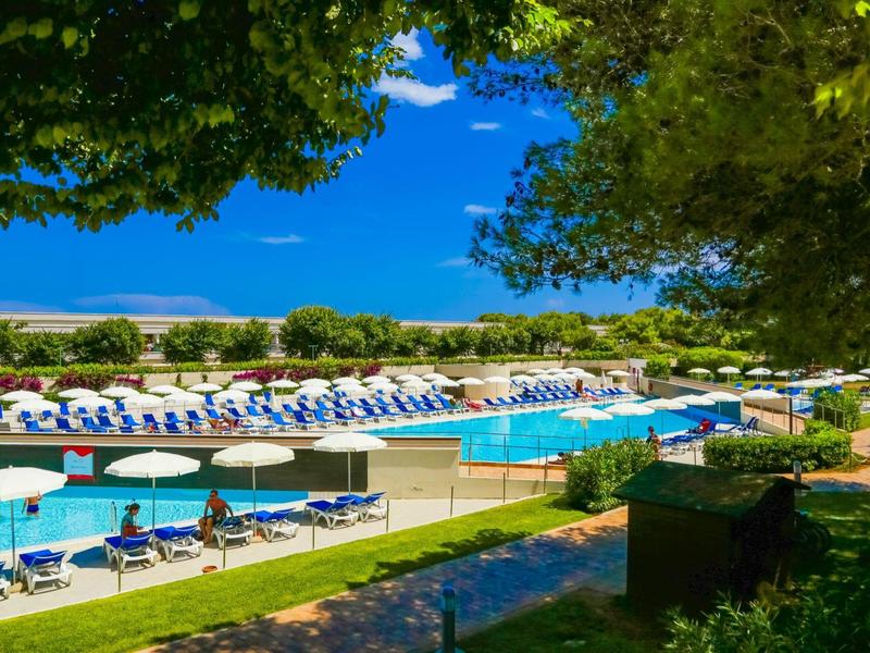 Grand piscine extérieure avec de nombreux parasols et transats dans un jardin d'hôtel sous un ciel bleu.