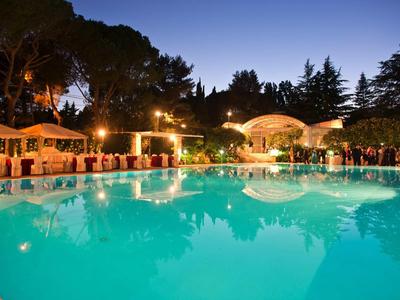 Beleuchteter Pool in Freien mit Stühlen und Tischen, umgeben von Bäumen bei Dämmerung.