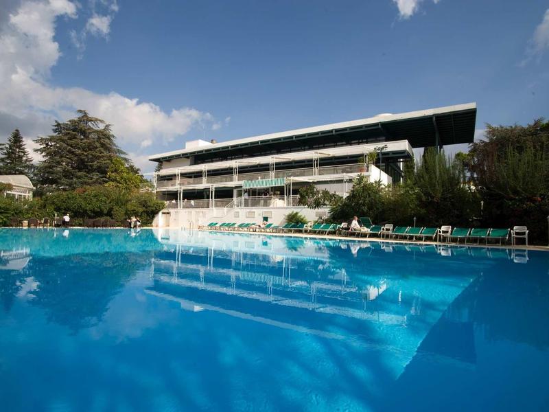 Großer Outdoor-Pool vor einem modernen Hotelgebäude mit klar blauem Himmel und grünen Bäumen.