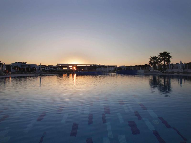 Calm pool at sunset with palm trees and buildings in the background.