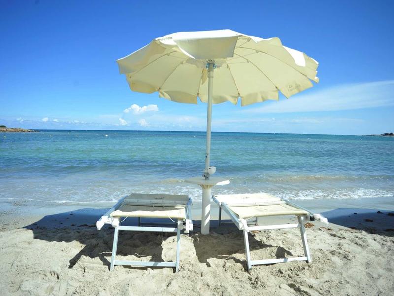 Two lounge chairs with a white umbrella on the sandy beach overlooking the sea.