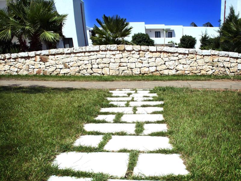 Stone path leads through green grass to white building with palm trees and stone wall.