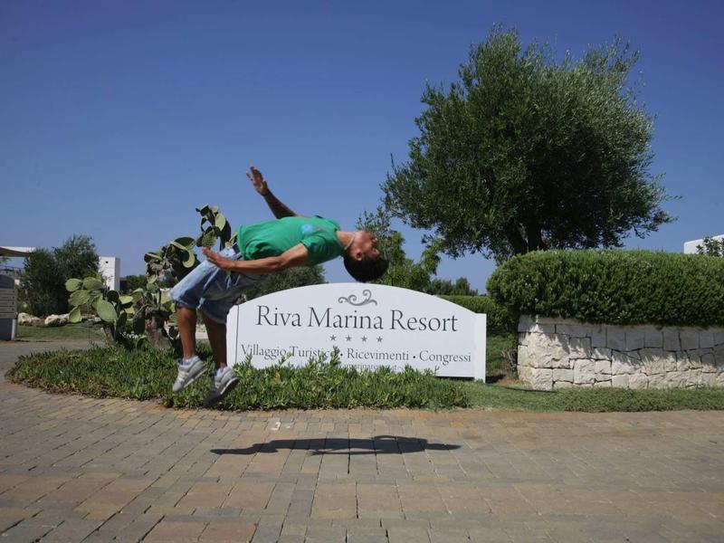 A man is jumping backward in front of a Riva Marina Resort sign under a clear blue sky.