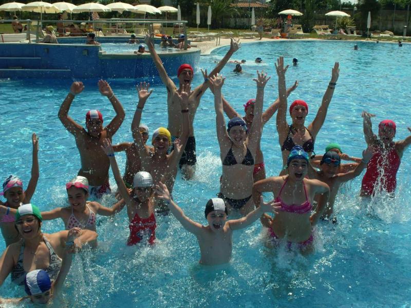 Children wearing colorful swim caps in a pool raising their arms and smiling.