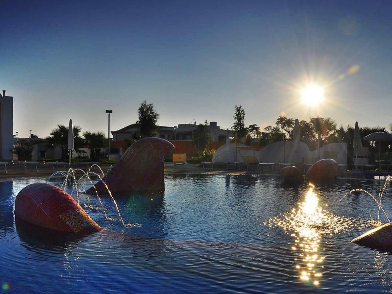 Man relaxing in pool at sunset with floating red spheres and houses in background.