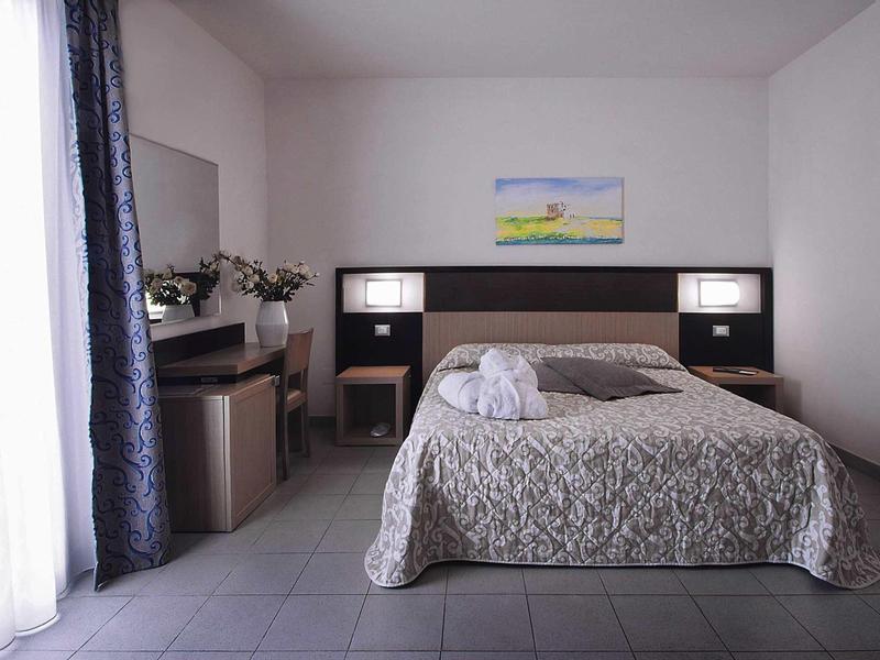 Bright hotel room with double bed, night lamps, table, chair, and curtain by the window.