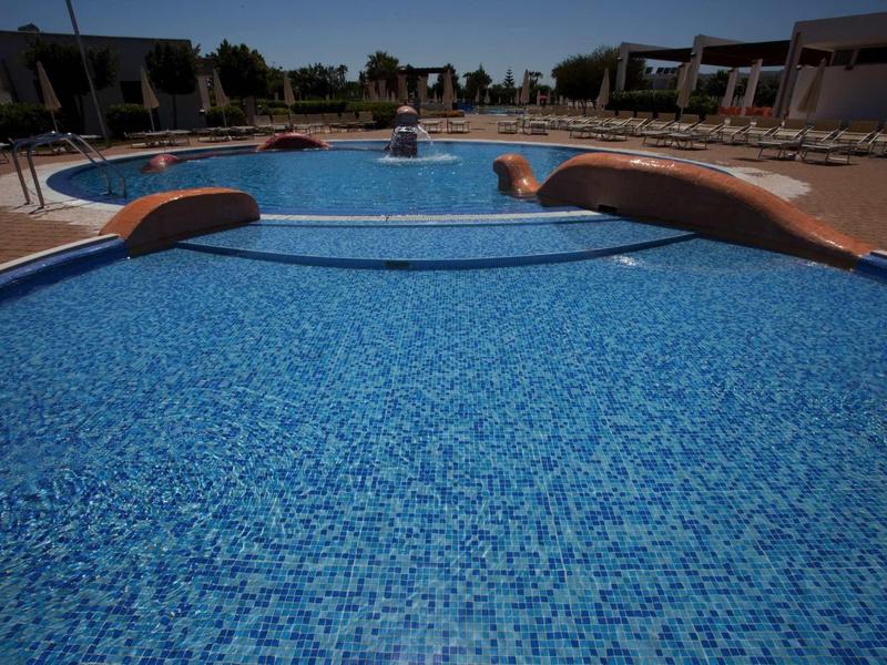 Large blue swimming pool with a bridge, surrounded by lounge chairs and buildings under clear sky.