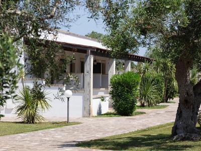 Weißes Haus mit Veranda, umgeben von grünen Bäumen, Sträuchern und einem gepflasterten Weg.