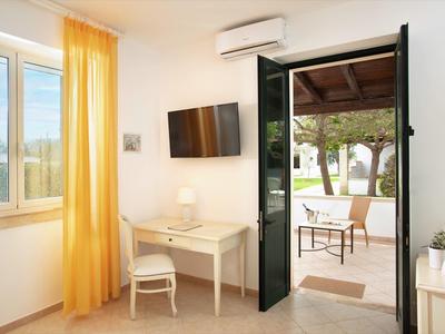 Helles Zimmer mit gelben Vorhängen, TV, Schreibtisch und offenem Tür zur Terrasse mit Gartenmobiliar.