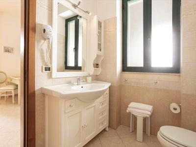 Helles Badezimmer mit weißem Waschbecken, großem Spiegel und Fenster, beige Fliesen und kleiner Hocker.