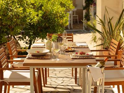 Tisch mit gedeckten Tellern und Gläsern, umgeben von Holzstühlen, draußen unter Baum und Sonne.