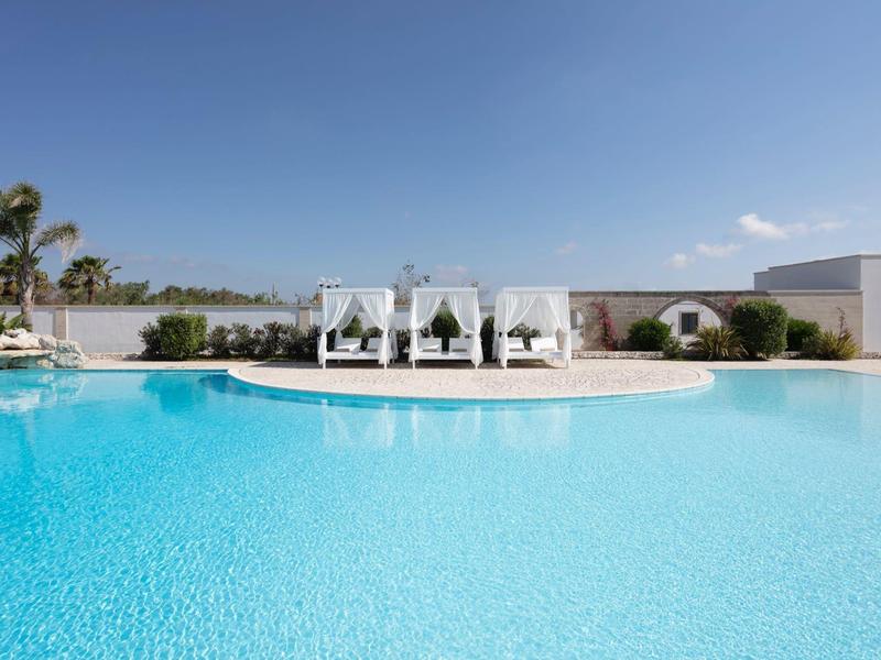 Großer, klarer Pool vor weißem Gebäude mit Sonnendächern und Palmen unter blauem Himmel.