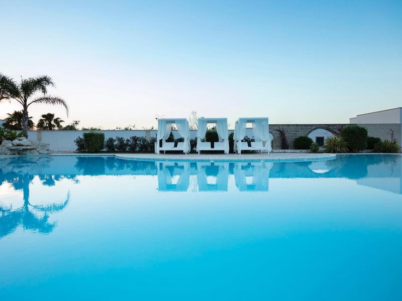 Infinity-Pool mit weißen Liegen und Palmen vor ruhigem Meer unter klarem Himmel.