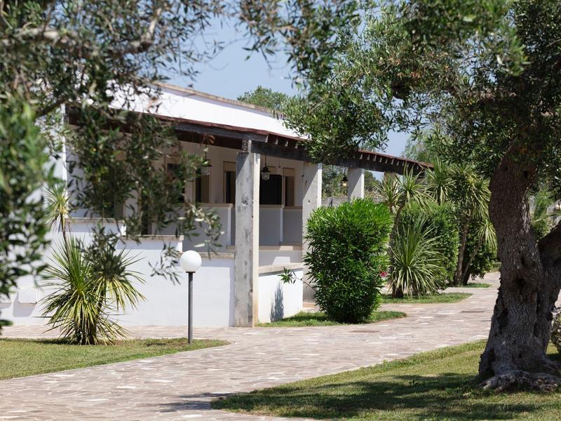 Weißes Haus mit Veranda, umgeben von grünen Bäumen, Sträuchern und einem gepflasterten Weg.