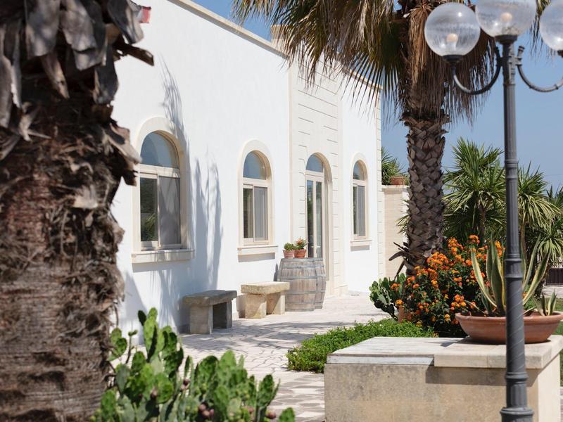 Weiße Hausfassade mit Bögen, Palmen, Pflanzen und schwarzer Laterne im sonnigen Garten.