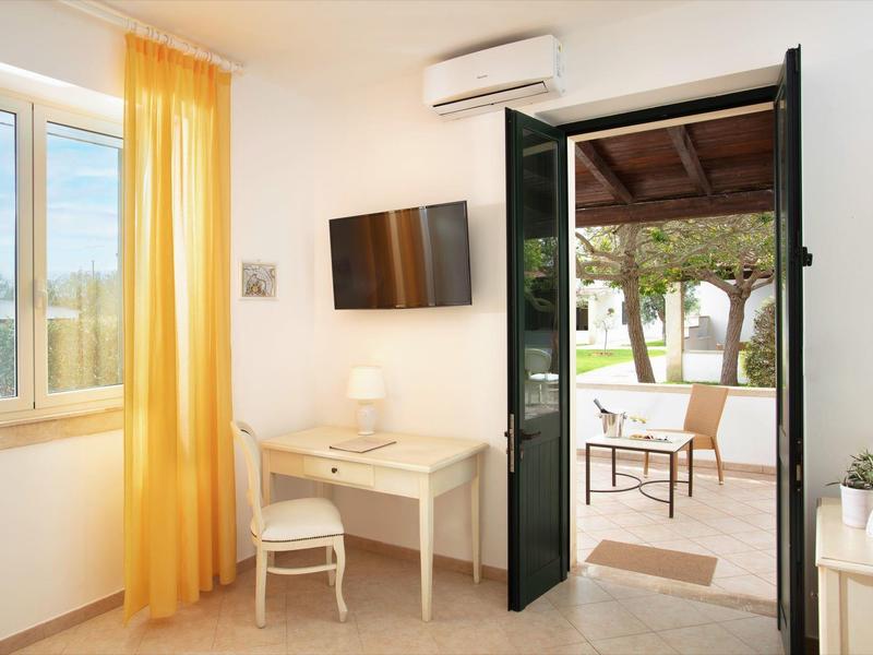 Helles Zimmer mit gelben Vorhängen, TV, Schreibtisch und offenem Tür zur Terrasse mit Gartenmobiliar.