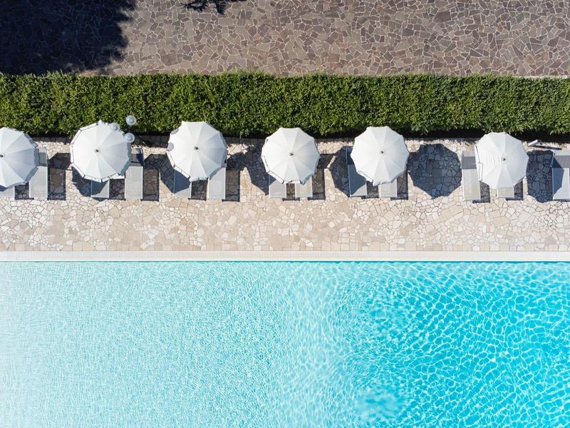 Dachterrasse mit weißen Sonnenschirmen am blauen Pool und grünem Heckenstreifen im Sonnenlicht.