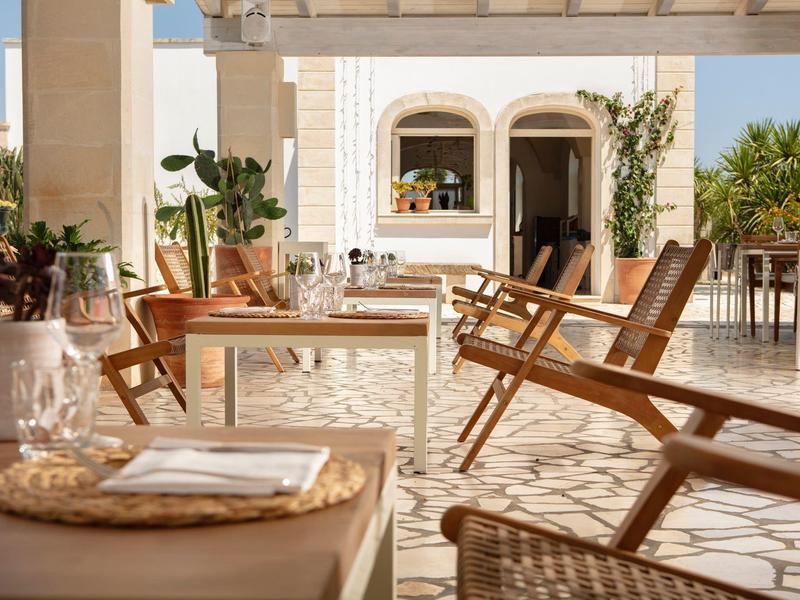 Mediterrane Terrasse mit Holzstühlen, Tischen, Pflanzen und heller Steinböden im Sonnenlicht.