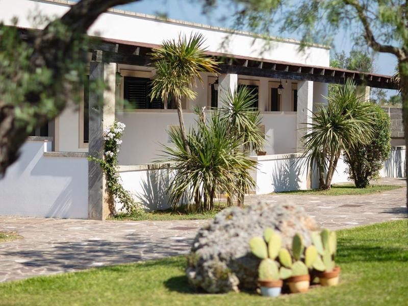 Ein weißes Gebäude mit Veranda und großen Pflanzen, umgeben von grünem Gras und Steinen.