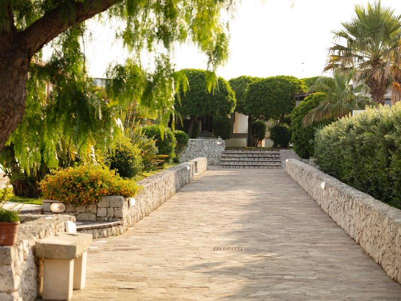 Breiter, sonniger Gartenweg mit Steinmauern, Büschen und großen Bäumen an Seiten.
