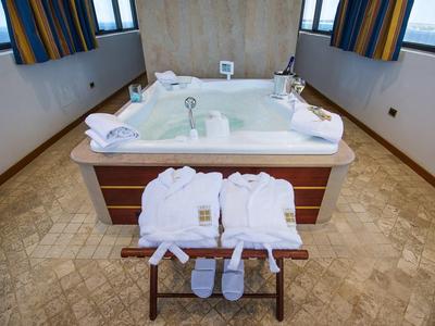 Ein großes, quadratisches Whirlpool-Bad mit weißen Bademänteln und Slippern auf einem Holzhocker davor.