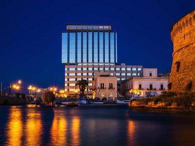 Beleuchtetes Hotelgebäude und historischer Turm am Wasser bei Nacht mit Spiegelungen.