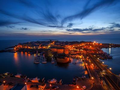 Hafenstadt bei Dämmerung mit beleuchteten Gebäuden und ruhigem Wasser unter blauem Himmel.