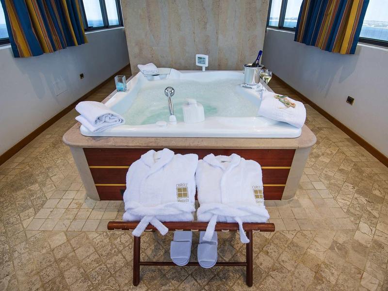 Ein großes, quadratisches Whirlpool-Bad mit weißen Bademänteln und Slippern auf einem Holzhocker davor.