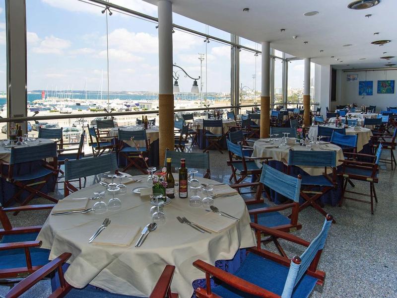 Hell eingerichteter Speisesaal mit blauen Stühlen und Blick auf den Hafen durch große Fenster.