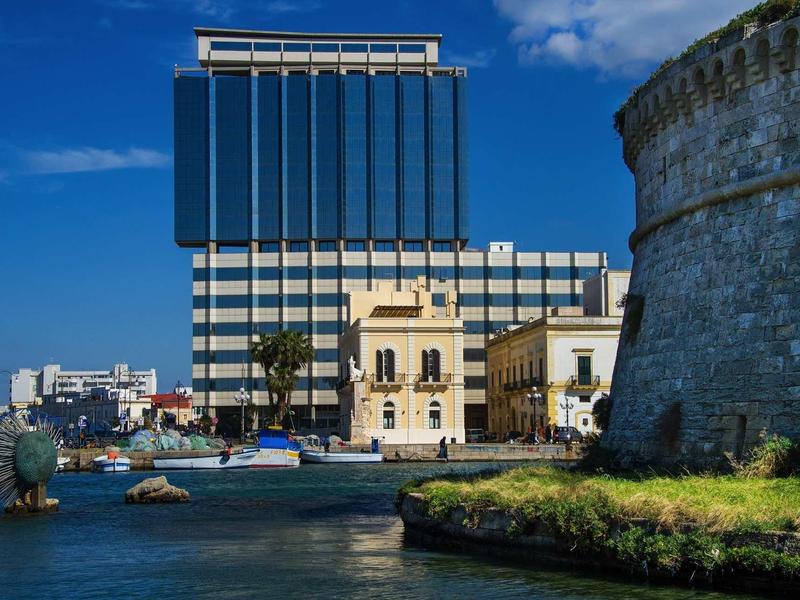 Modernes Gebäude und alte Festungsmauer am Wasser unter strahlend blauem Himmel.