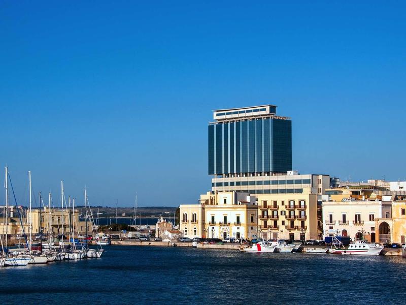 Hafen mit Segelbooten, blauem Himmel und modernen sowie historischen Gebäuden am Wasser.