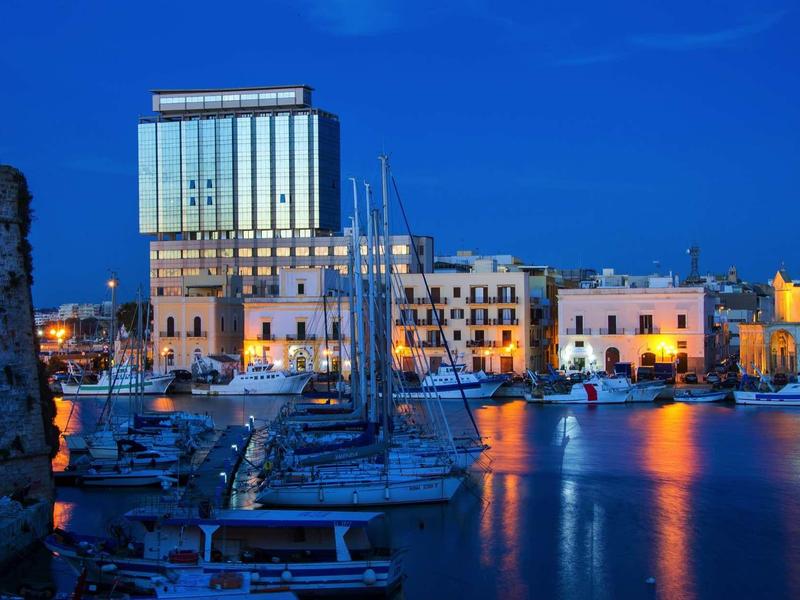 Hafen mit Segelbooten vor beleuchteten Gebäuden und blauem Nachthimmel.