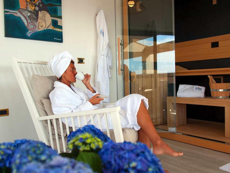 Frau in weißem Bademantel und Turban sitzt entspannt auf weißem Schaukelstuhl auf Balkon.