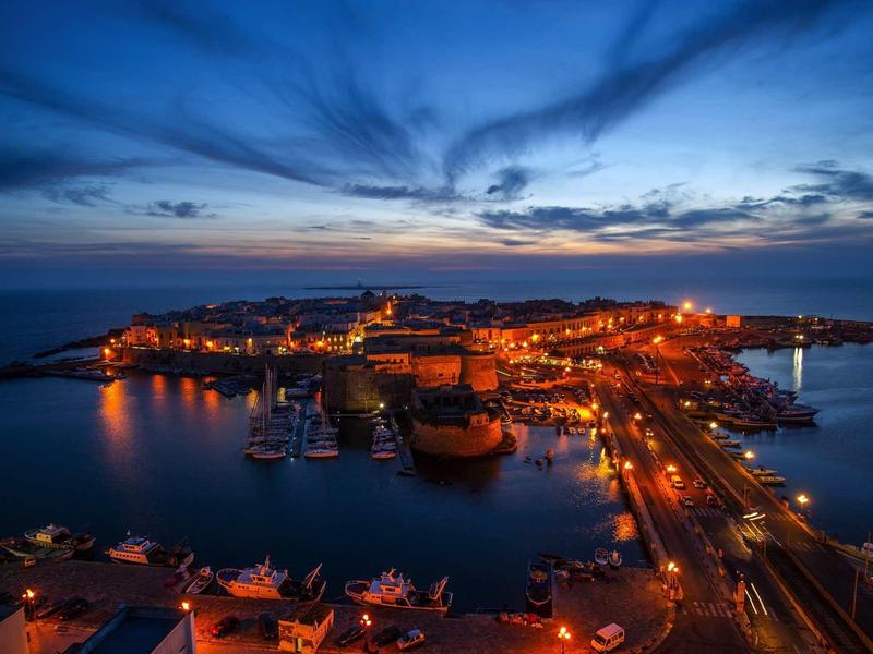Hafenstadt bei Dämmerung mit beleuchteten Gebäuden und ruhigem Wasser unter blauem Himmel.