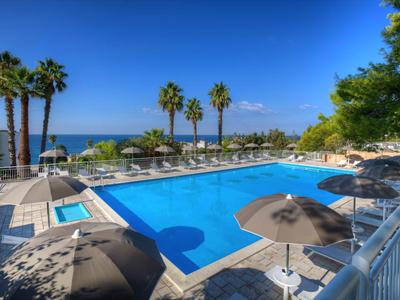 Großer Außenpool mit blauer Wasseroberfläche, umgeben von Liegestühlen, Sonnenschirmen und Palmen am Meer.