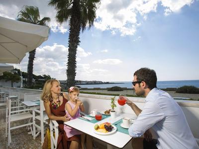 Familie sitzt draußen an einem weißen Tisch mit Blick auf Meer und Palmen bei Tageslicht.