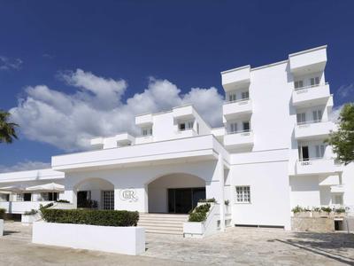 Modernes weißes Gebäude mit mehreren Stockwerken, Balkonen und Palmen vor blauem Himmel.